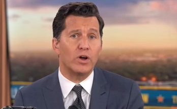 man in suit and tie speaking on TV news set with cityscape backdrop