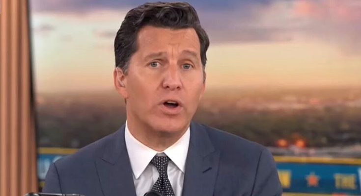 man in suit and tie speaking on TV news set with cityscape backdrop