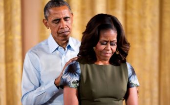 barack and Michelle Obama stand together, hands on shoulders, looking down with solemn