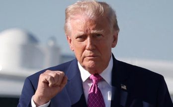 former President Donald Trump raises his fist outdoors near a large white aircraft