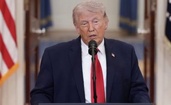 former President Donald Trump speaking at podium with American flag behind him wearing