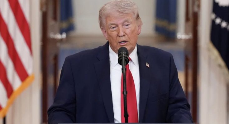 former President Donald Trump speaking at podium with American flag behind him wearing
