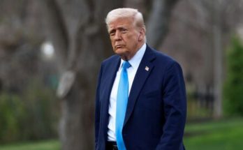 former President Donald Trump in navy suit and blue tie standing outdoors near trees