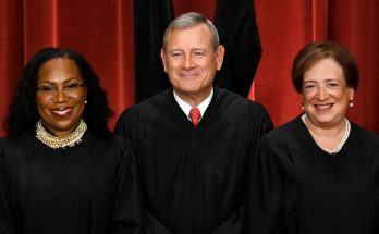 three Supreme Court Justices smiling in black robes against a red curtain backdrop