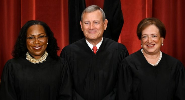 three Supreme Court Justices smiling in black robes against a red curtain backdrop