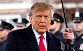 donald Trump holding black umbrella, speaking outdoors with police officers in background