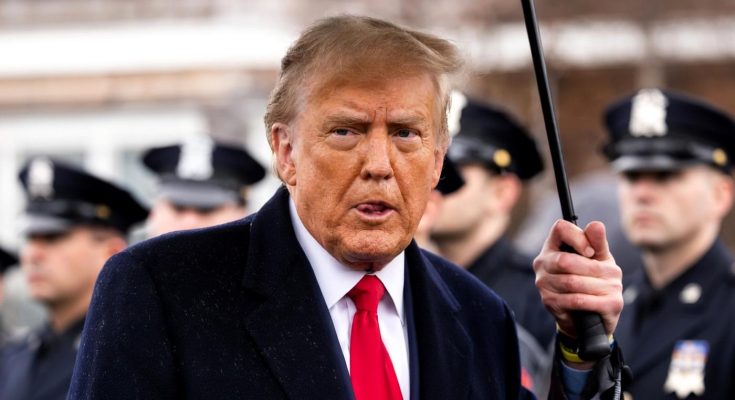 donald Trump holding black umbrella, speaking outdoors with police officers in background