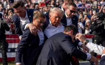 security personnel protect a public figure at outdoor event, crowd in background