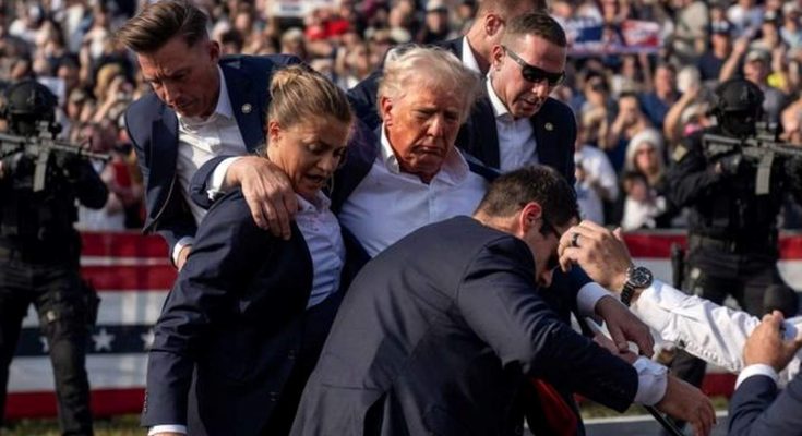 security personnel protect a public figure at outdoor event, crowd in background