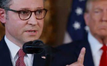 man in suit and glasses speaking at podium with American flags behind him, another man