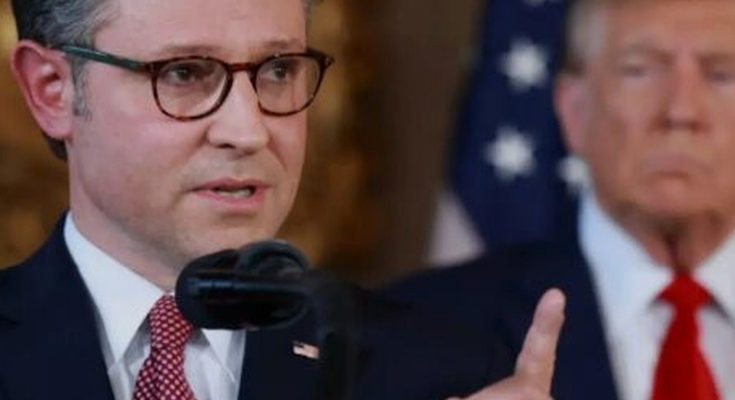 man in suit and glasses speaking at podium with American flags behind him, another man