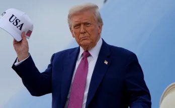 donald Trump holding a white 'USA' cap with American flag patch, standing on an airplane