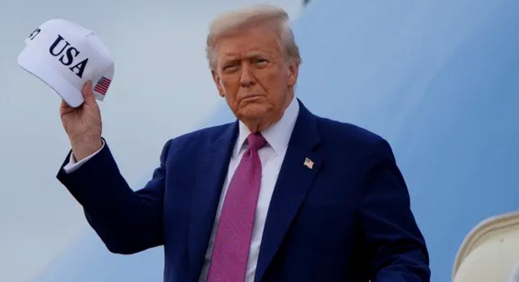 donald Trump holding a white 'USA' cap with American flag patch, standing on an airplane