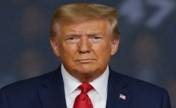 donald Trump in navy suit with red tie and American flag pin, serious expression against