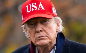 donald Trump wearing a red USA baseball cap with American flag patch, outdoors with