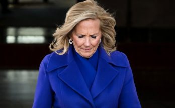 woman with blonde hair in a blue coat looking down with a somber expression