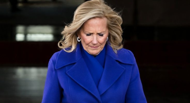 woman with blonde hair in a blue coat looking down with a somber expression