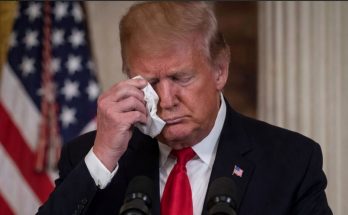 former President Donald Trump wiping his eye with a tissue at a podium with an American