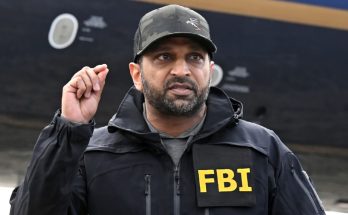 FBI agent in black jacket and cap gestures while standing in front of aircraft