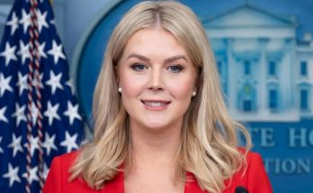 woman in red blazer smiles at camera in White House press briefing room with American