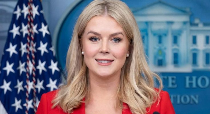 woman in red blazer smiles at camera in White House press briefing room with American
