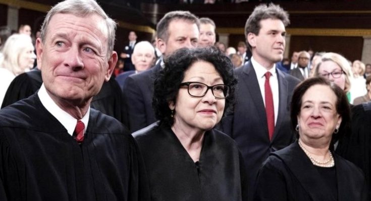 three Supreme Court justices in black robes stand together in a formal courtroom setting