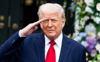 donald Trump saluting in dark suit and red tie with floral arrangements behind him