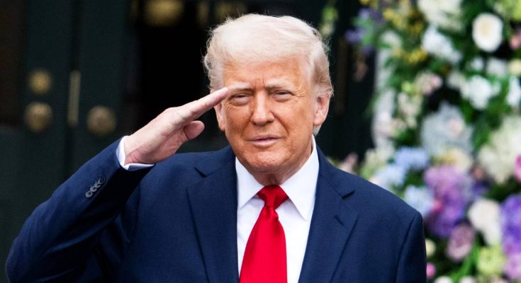donald Trump saluting in dark suit and red tie with floral arrangements behind him