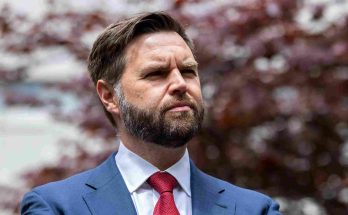 man with beard in blue suit and red tie standing outdoors with blurred red foliage