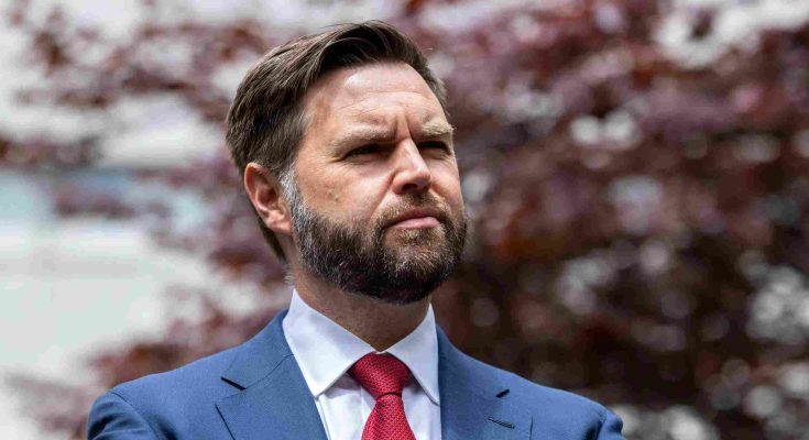 man with beard in blue suit and red tie standing outdoors with blurred red foliage
