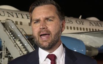 man in suit and red tie speaking in front of a United States of America airplane at night