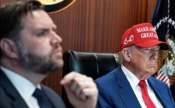 two men in a meeting room, one wearing a red hat with slogan