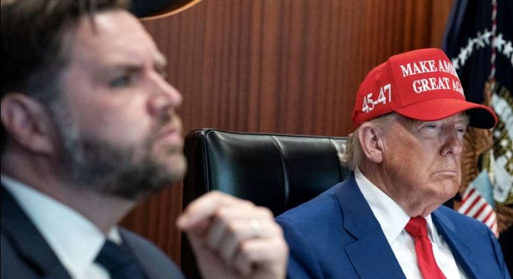 two men in a meeting room, one wearing a red hat with slogan